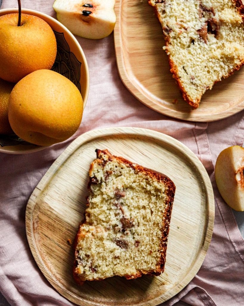 A wooden plate with a slice of pound cake sits on a pink cloth next to a bowl of whole Asian pears and another plate with a partially eaten slice of cake. Some pear slices are scattered nearby.