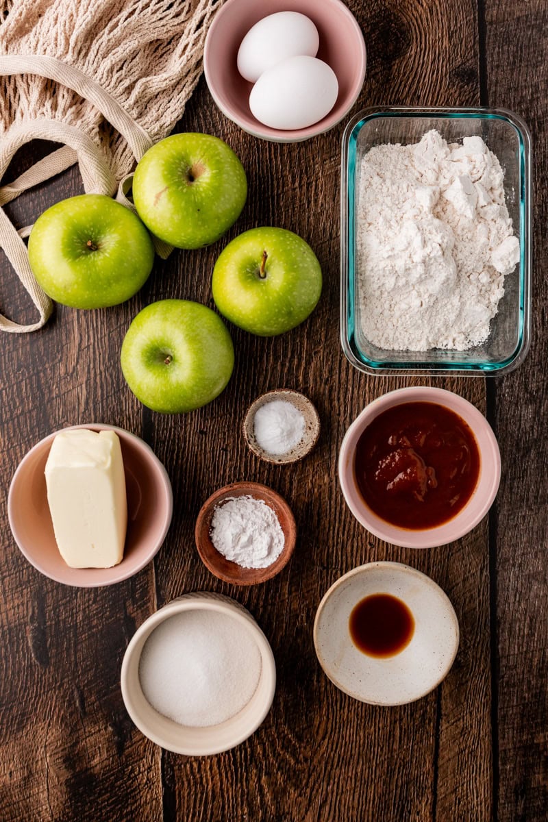 A flat lay of baking ingredients for apple pumpkin bread on a wooden surface: three green apples, two eggs, a glass container of flour, a bowl of butter, a small bowl of sugar, baking powder, cinnamon, salt, and vanilla extract. A cloth bag is also visible.