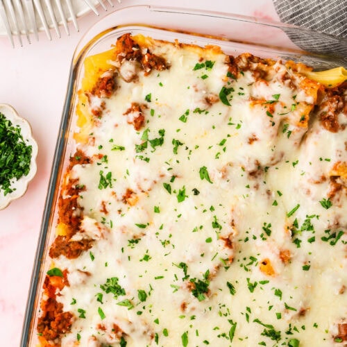 A casserole dish filled with baked lasagna topped with melted cheese and chopped parsley—proof that not all fails of pasta end badly—sits next to a gray napkin, a small dish of fresh herbs, and a plate with four forks.