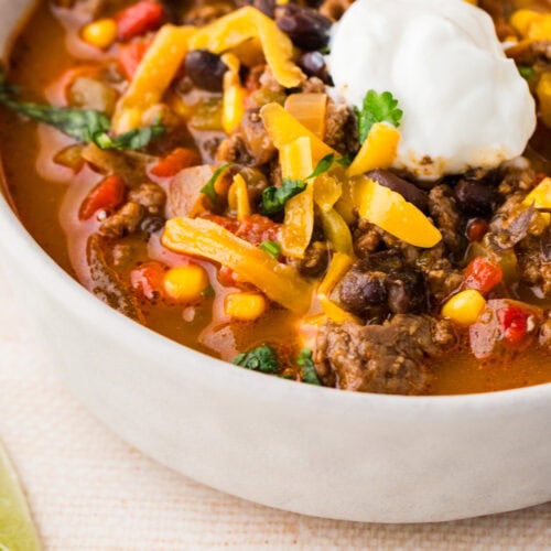 A close-up of a bowl of spicy beef soup taco topped with shredded cheddar cheese, a dollop of sour cream, and fresh cilantro. The soup contains ground beef, black beans, corn, and tomatoes. A tortilla chip rests on the bowl’s edge.