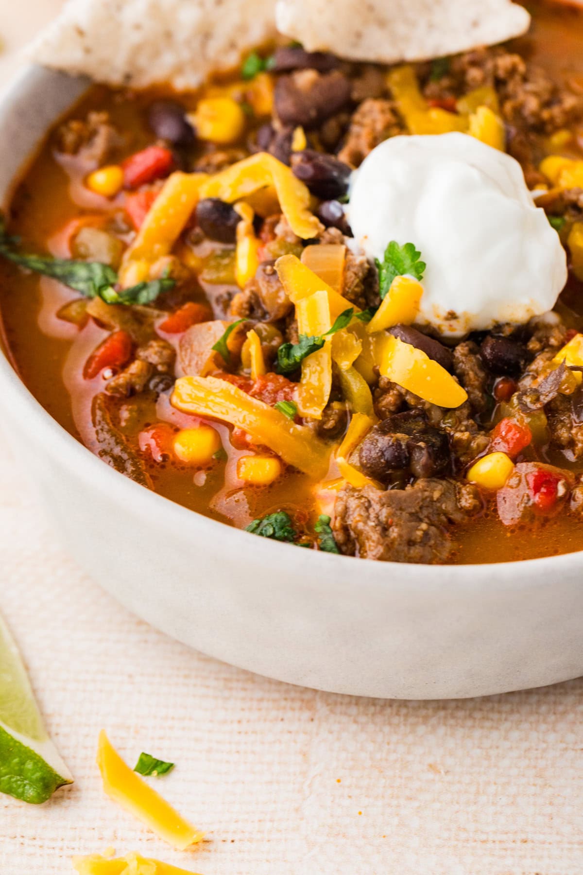 A close-up of a bowl of spicy beef soup taco topped with shredded cheddar cheese, a dollop of sour cream, and fresh cilantro. The soup contains ground beef, black beans, corn, and tomatoes. A tortilla chip rests on the bowl’s edge.