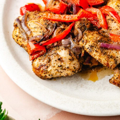 A plate of seasoned Italian baked chicken thighs topped with slices of red bell peppers and red onions, garnished with herbs. A branch of fresh parsley is partially visible in the corner, all set on a light pink surface.
