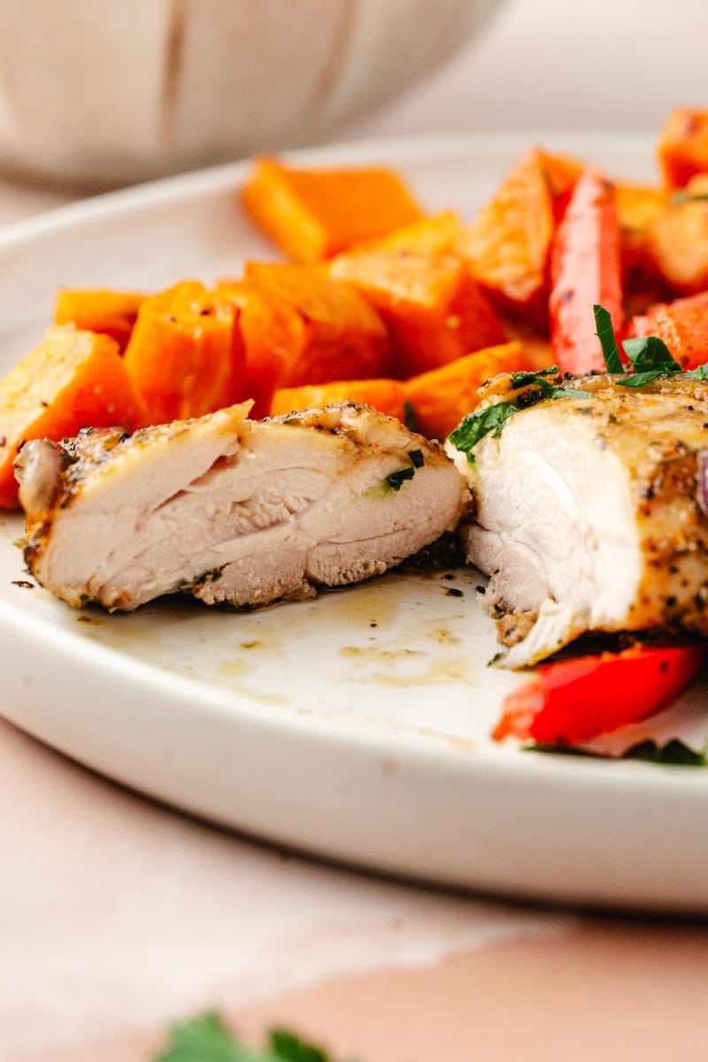 A close-up of a plate with sliced, cooked chicken breast seasoned with Italian herbs. In the background, roasted orange vegetables, possibly sweet potatoes or carrots, and red bell peppers complement the dish. Reminiscent of a hearty Italian baked chicken thigh meal, it's garnished with fresh green herbs.