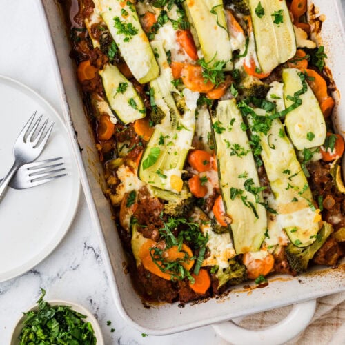 A casserole dish filled with sliced zucchini, carrots, broccoli, and melted cheese creates an easy zucchini lasagna. Fresh herbs are sprinkled on top. Nearby are empty white plates with forks and small bowls containing chopped herbs, all resting on a marble surface.