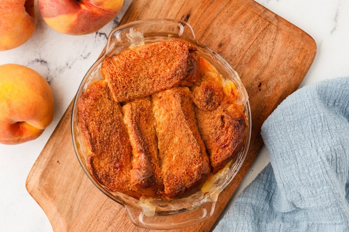 A glass dish of mini peach cobbler with golden brown crust sits on a wooden board, surrounded by fresh peaches and a light blue cloth.