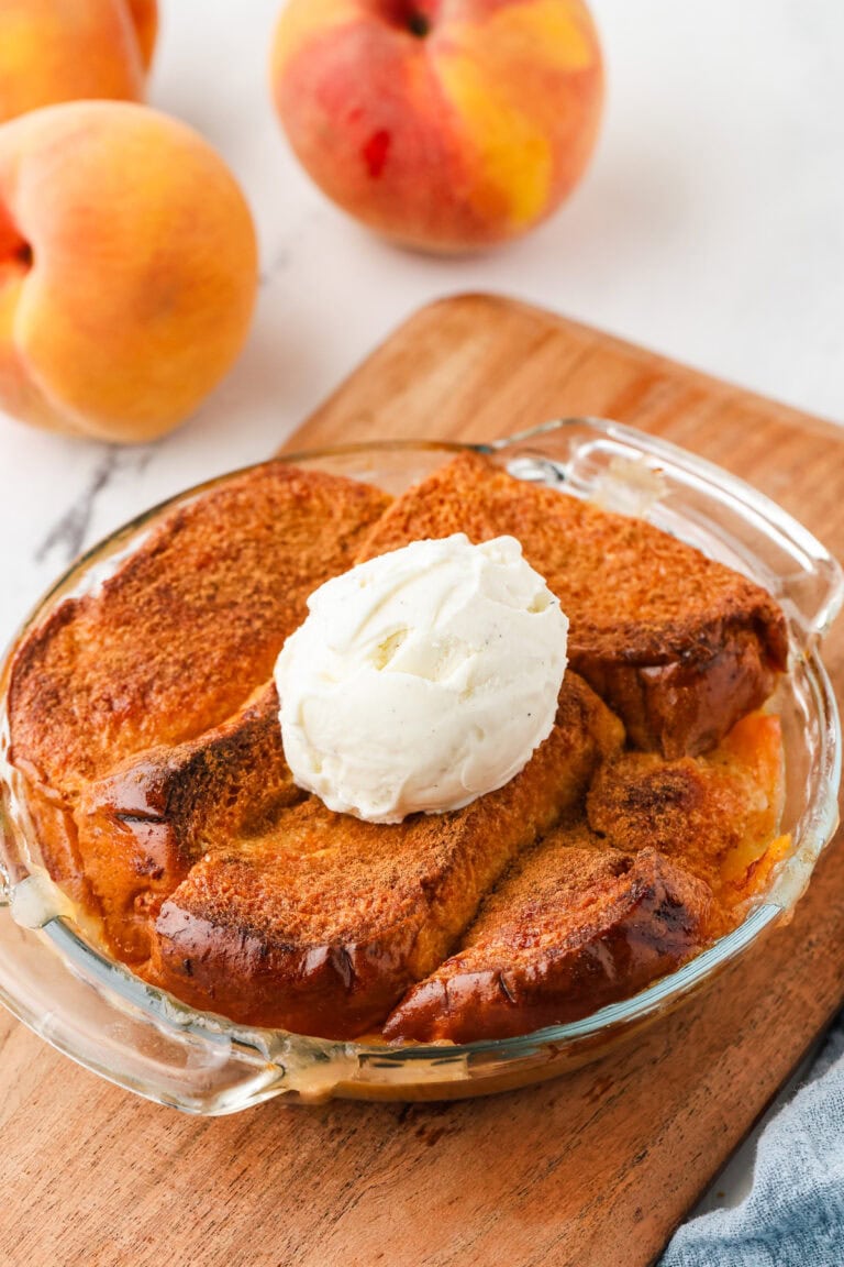 A mini peach cobbler, baked to golden perfection in a glass dish and topped with a scoop of vanilla ice cream, sits invitingly on a wooden board. Fresh peaches are arranged in the background.