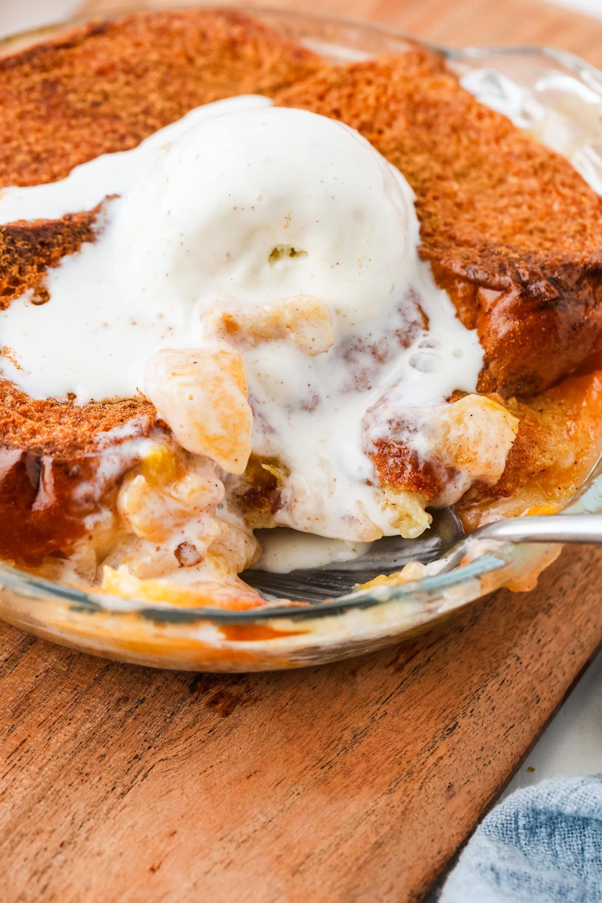 A close-up of mini peach cobbler topped with melting vanilla ice cream in a glass dish, with a spoon scooping out a portion. The golden-brown cobbler reveals juicy peaches beneath its crisp topping.