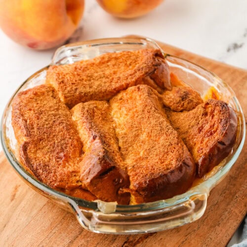 A glass baking dish filled with baked bread pudding sits on a wooden board, reminiscent of a comforting mini peach cobbler, with fresh peaches in the background.