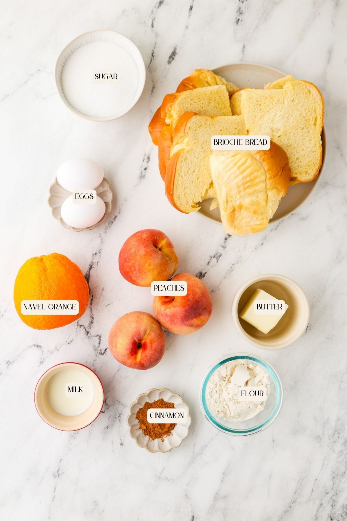 Slices of brioche bread, eggs, butter, peaches, a navel orange, sugar, milk, cinnamon, and flour are arranged on a marble surface—each labeled with its name—ready to come together for a delightful mini peach cobbler.