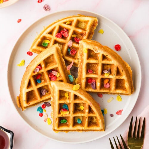 A white plate holds four golden Fruity Pebble Waffles quarters topped with colorful cereal pieces. More Fruity Pebbles are scattered on the table beside forks, a napkin, syrup, and extra waffle pieces.