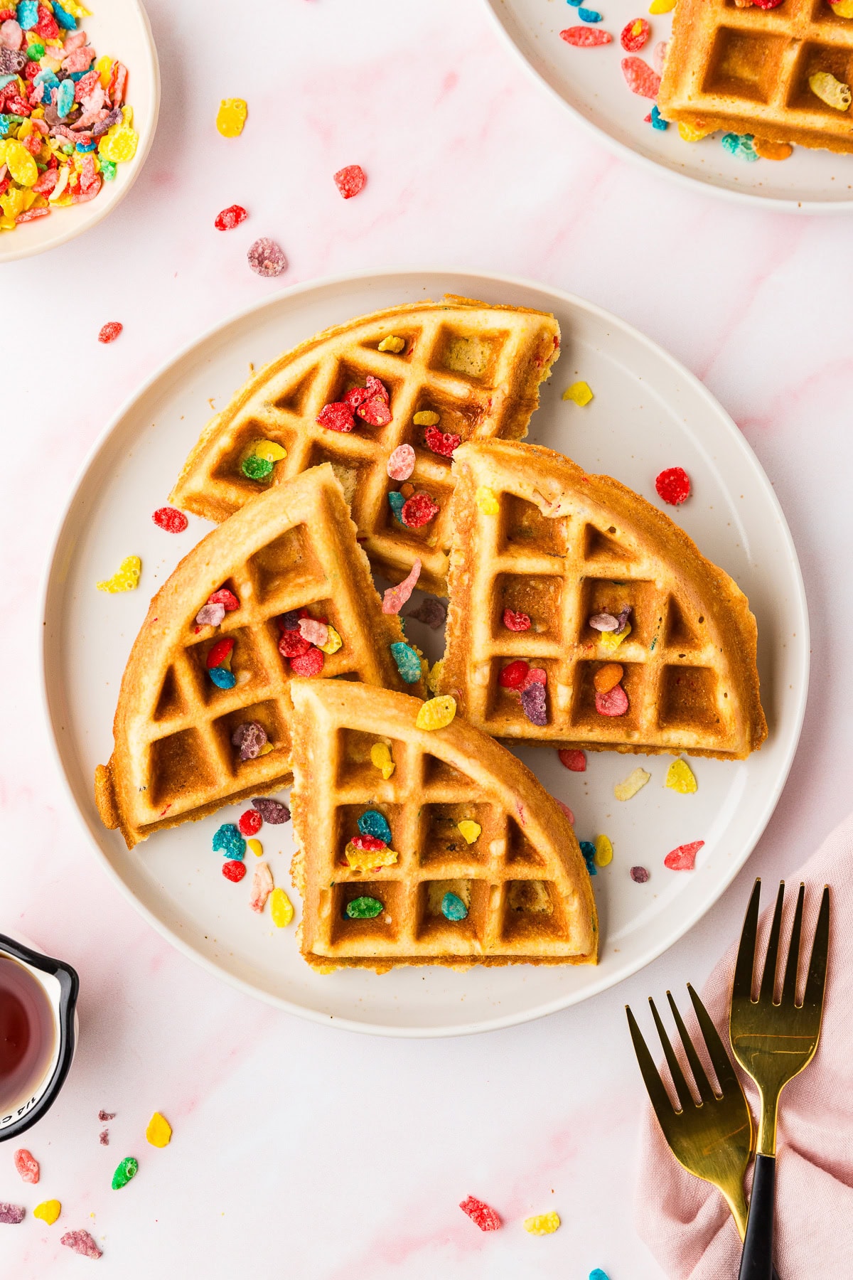 A white plate holds four golden Fruity Pebble Waffles quarters topped with colorful cereal pieces. More Fruity Pebbles are scattered on the table beside forks, a napkin, syrup, and extra waffle pieces.