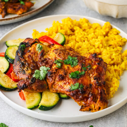 A plate of seasoned Greek yogurt chicken garnished with cilantro, served with sautéed zucchini and red bell peppers, alongside yellow rice. A bowl of rice and another plate of chicken are in the background.