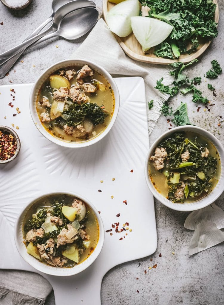 3 bowls of soup with kale and potatoes with spoons on a white board