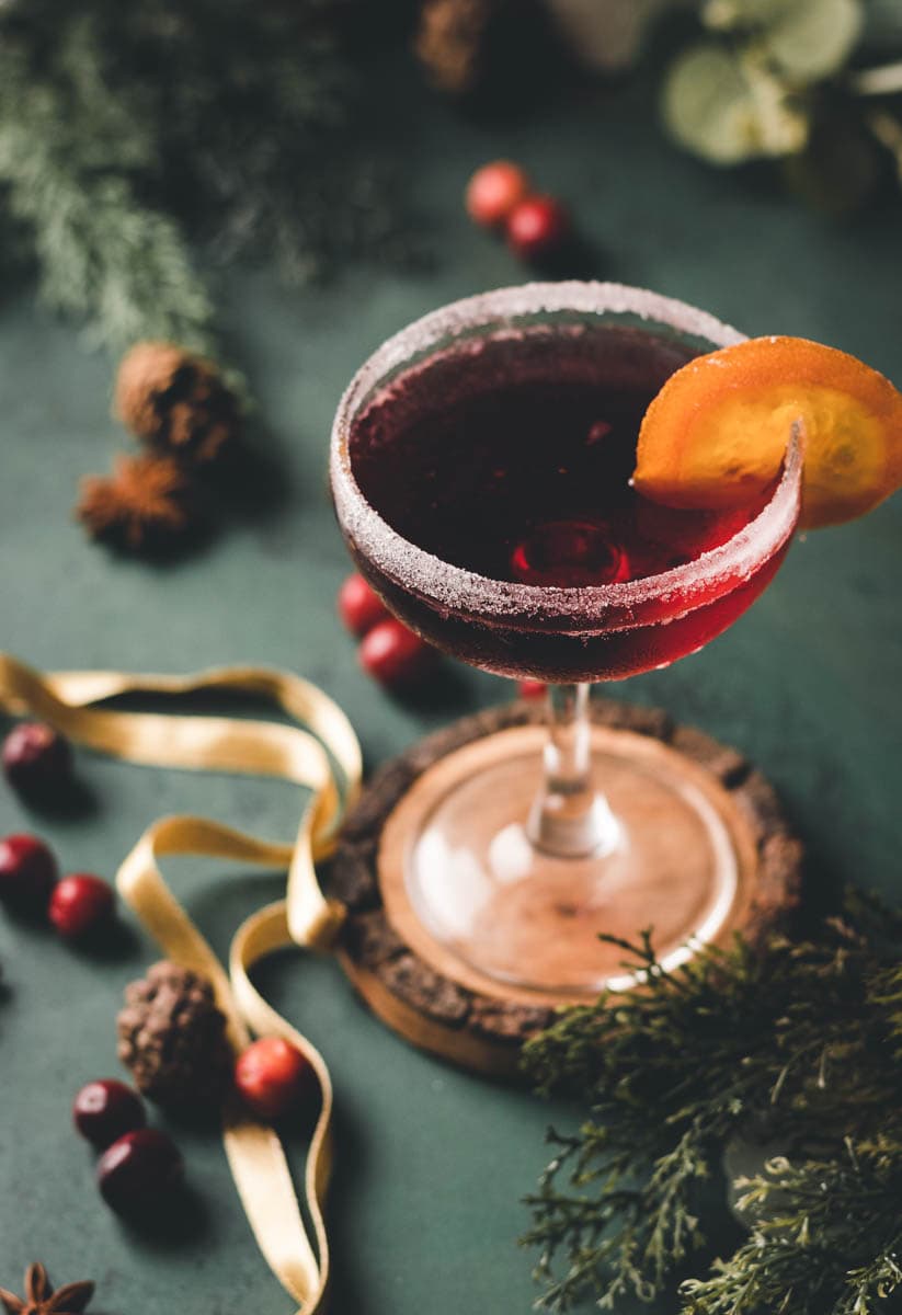 cranberry mocktail with an orange garnish in a coupe glass on top of a wood coaster, sitting on a table with pine cones, pine needles and cranberries.