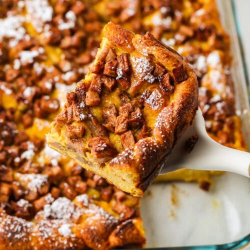 A spatula holds a square piece of pumpkin French toast casserole topped with diced cinnamon apples, above a glass baking dish with more pieces inside.