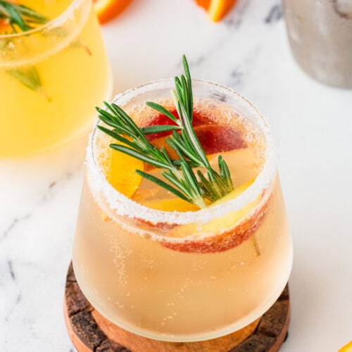 A white sangria mocktail garnished with orange slices and a sprig of rosemary sits on a wooden coaster, surrounded by fresh orange wedges and another drink in the background on a white surface.