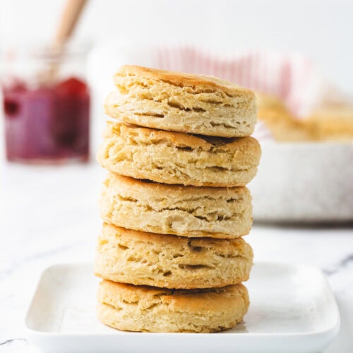 A stack of four golden-brown, gluten-free buttermilk biscuits sits on a square white plate. In the background, a jar with likely jam and a spreading knife peeks out, all set against a white marble surface.