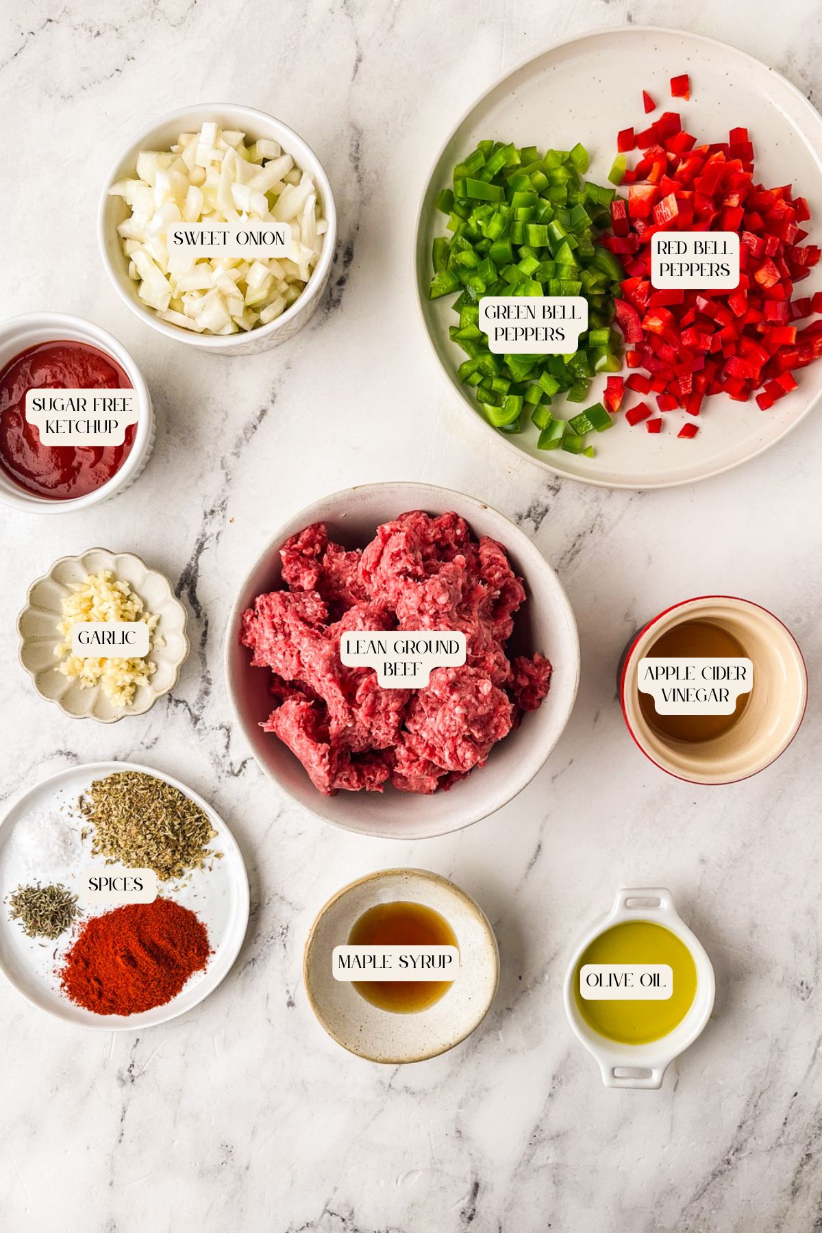 Bowls of chopped sweet onion, bell peppers, lean ground beef, sliced garlic, apple cider vinegar, olive oil, maple syrup, sugar-free ketchup, and a mix of spices ready for making sugar free sloppy joe's on a marble surface.