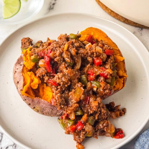A baked sweet potato topped with sugar free sloppy joe's mixture of ground beef, bell peppers, onions, and sauce sits on a white plate. A glass of water with lime slices is in the background.