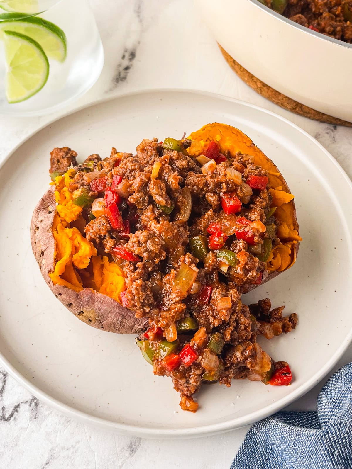 A baked sweet potato topped with sugar free sloppy joe's mixture of ground beef, bell peppers, onions, and sauce sits on a white plate. A glass of water with lime slices is in the background.