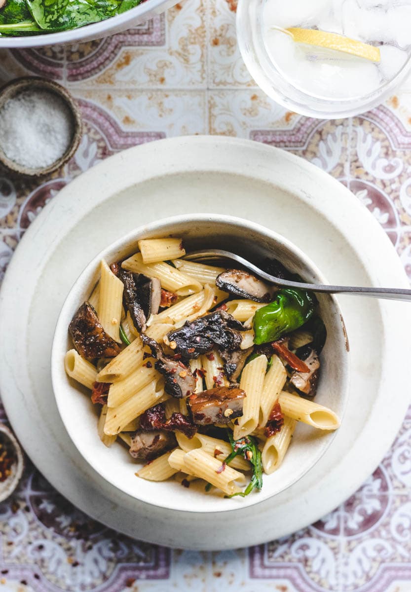 penne pasta in a white bowl with pepper flakes , portobello mushrooms and spinach.