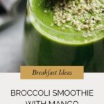 A green broccoli smoothie in a glass, topped with seeds and yellow edible flowers. Text overlay reads: Breakfast Ideas. Broccoli Smoothie with Mango. whensaltyandsweetunite.com.