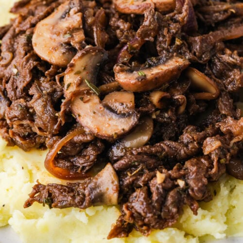 Shredded beef and sliced mushrooms in a rich brown sauce, inspired by a classic shaved steak recipe, served over creamy mashed potatoes on a white plate.