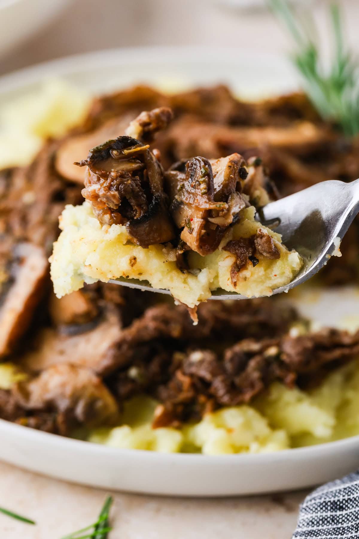A close-up of a fork holding mashed potatoes topped with tender beef and mushrooms, with more beef and mashed potatoes on a plate in the background—perfect for trying out your favorite shaved steak recipe.