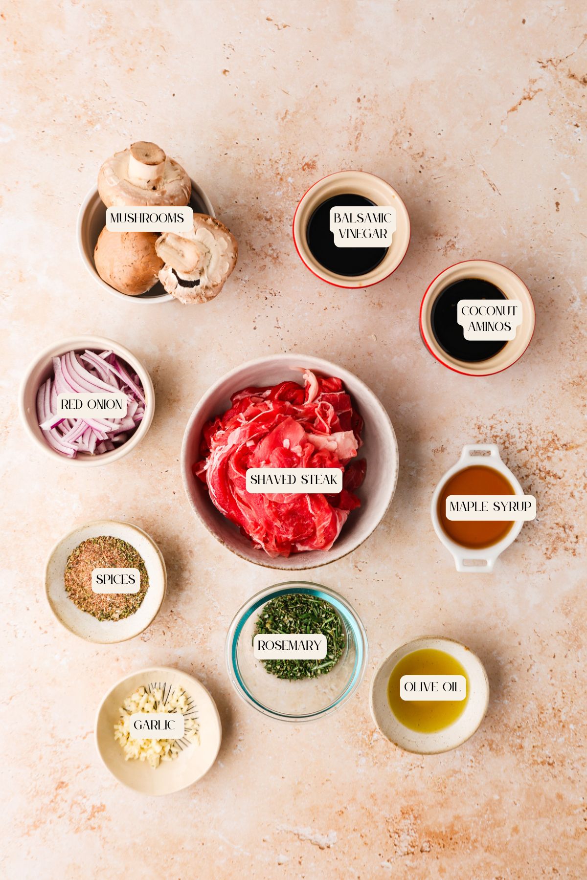 Top-down view of bowls with labeled ingredients for a delicious shaved steak recipe: mushrooms, red onion, spices, garlic, rosemary, olive oil, maple syrup, coconut aminos, and balsamic vinegar arranged on a light surface.