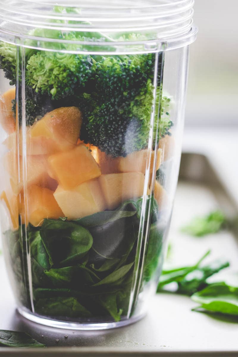 A clear blender cup filled with fresh spinach leaves, cubed cantaloupe, and broccoli florets sits on a white surface with scattered greens nearby, ready to be blended into a nutritious broccoli smoothie.