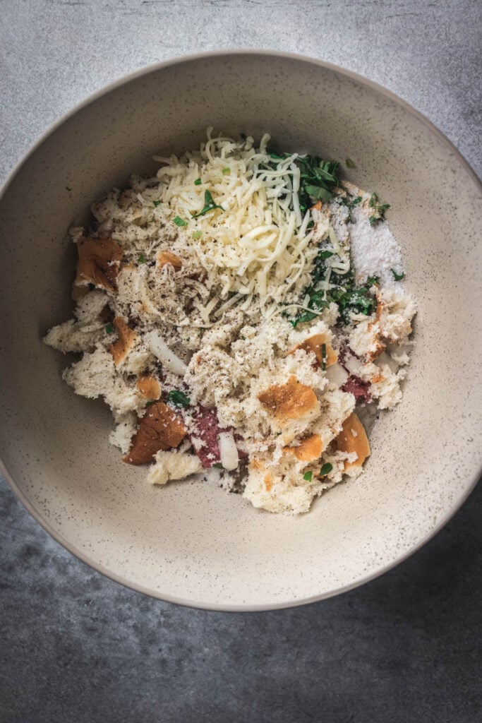 A bowl containing a mixture of torn bread, grated cheese, chopped herbs, salt, and pepper—perfect for making Hawaiian roll meatball sliders—sits ready to be combined on a gray surface.