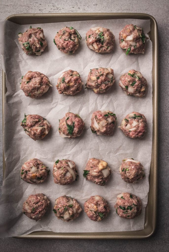 Twenty uncooked meatballs with visible herbs and onions are evenly spaced on a parchment-lined baking sheet, ready to be cooked for flavorful Hawaiian roll meatball sliders.