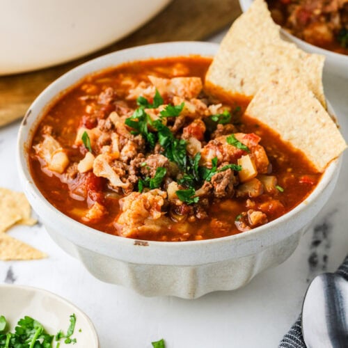 A bowl of cauliflower chili with ground meat, topped with tortilla chips and cilantro.