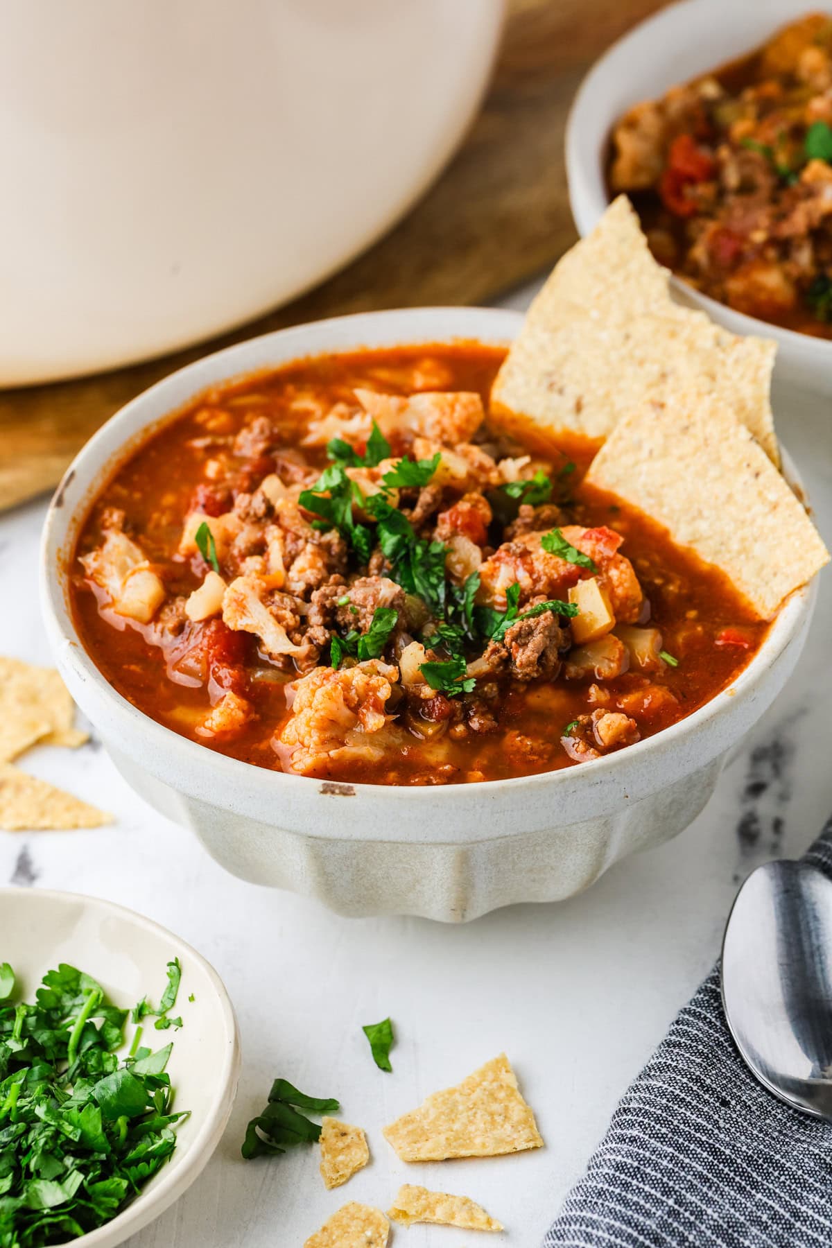 A bowl of cauliflower chili with ground meat, topped with tortilla chips and cilantro.