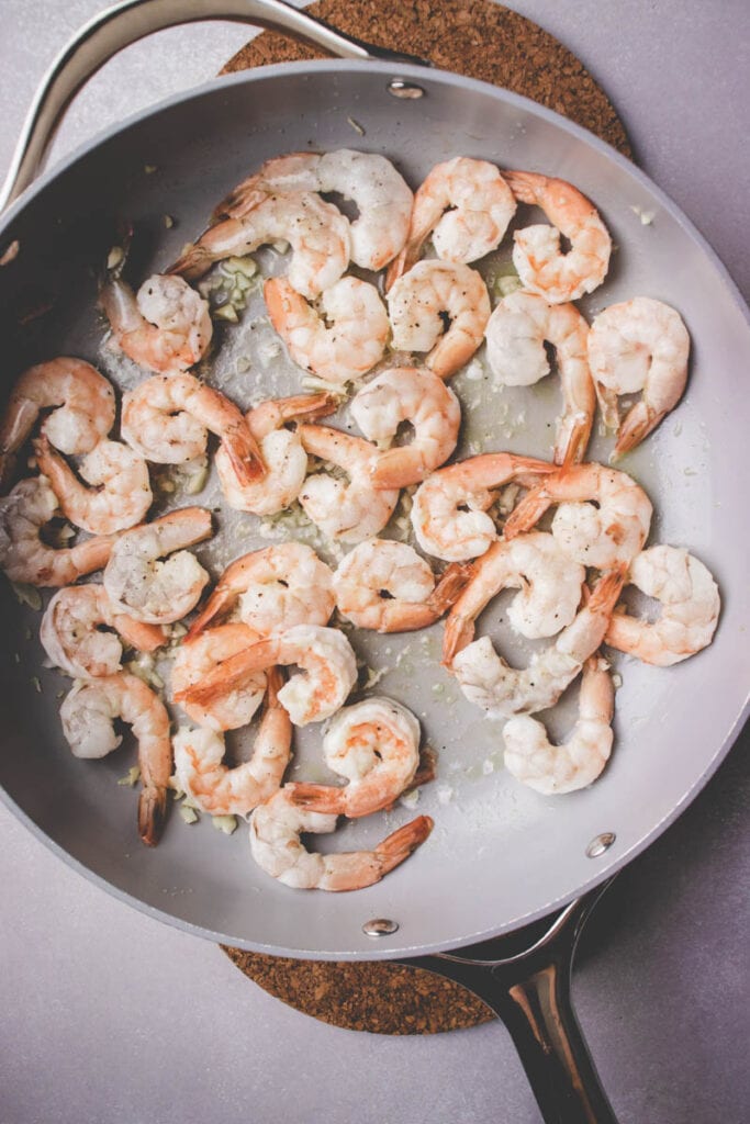 A silver skillet filled with cooked shrimp sautéing in oil and minced garlic, perfect as a topping for shrimp and zucchini risotto, resting on a round cork trivet on a gray surface.