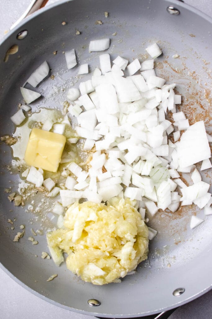 A frying pan containing chopped white onions, a pat of butter, and a mound of minced garlic—uncooked and ready to be sautéed for a flavorful shrimp and zucchini risotto.