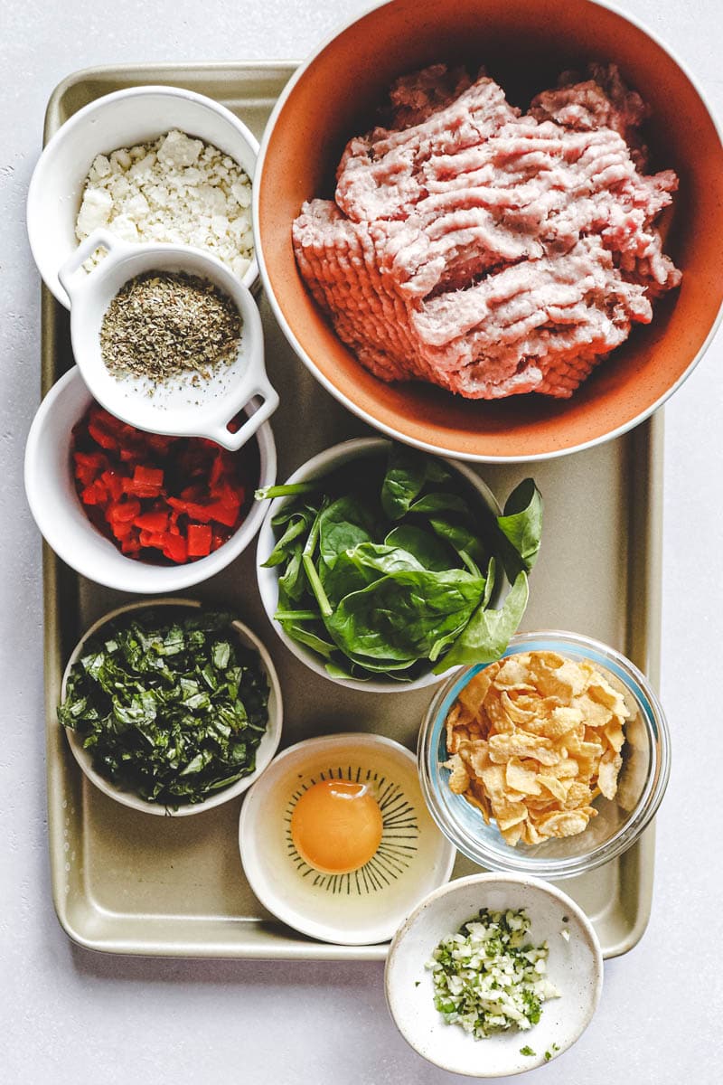 A tray with bowls containing ground meat for turkey basil burgers, crumbled cheese, dried herbs, chopped red peppers, fresh spinach leaves, chopped basil, a cracked egg, crushed cornflakes, and minced garlic.