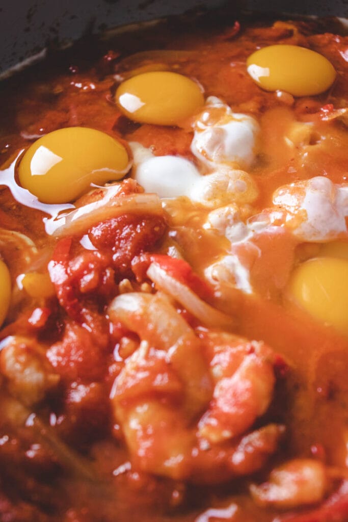 Four raw eggs are cracked over a tomato-based stew with visible chunks of shrimp and onions, ready to be cooked together in a pan.