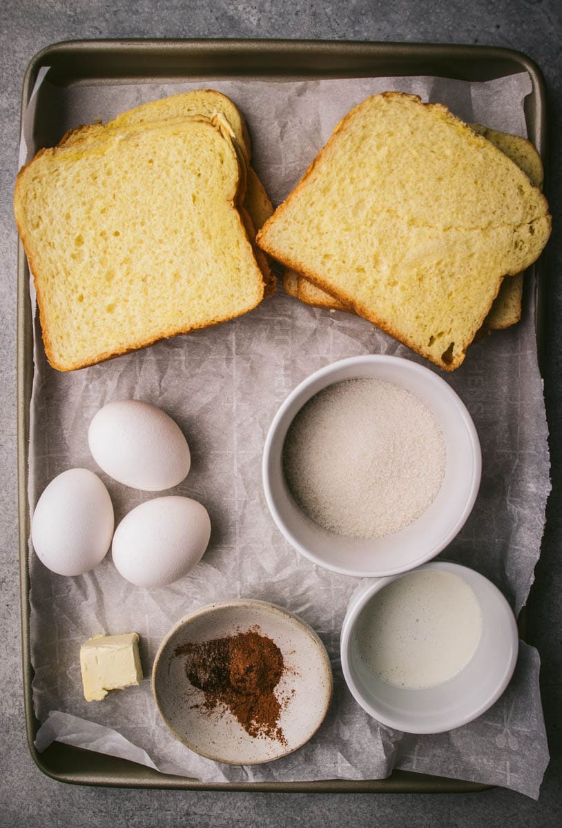 A baking tray with slices of bread, three eggs, a bowl of sugar, a bowl of milk, a pat of butter, and a dish with spices—all you need for delicious french toast without vanilla—arranged neatly on parchment paper.
