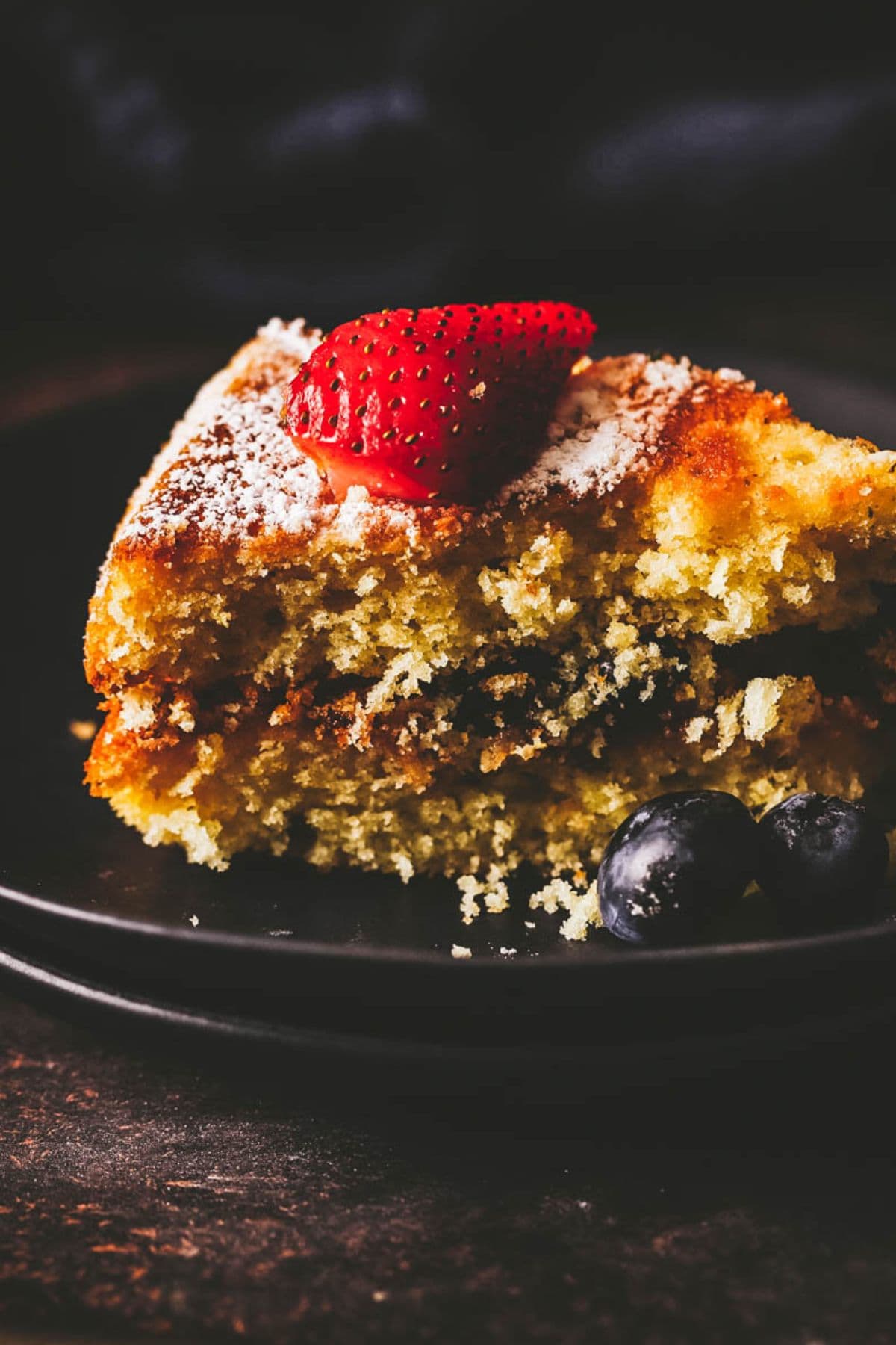 A slice of gluten free lemon cake topped with powdered sugar and a sliced strawberry, with two blueberries on the side, served on a black plate.
