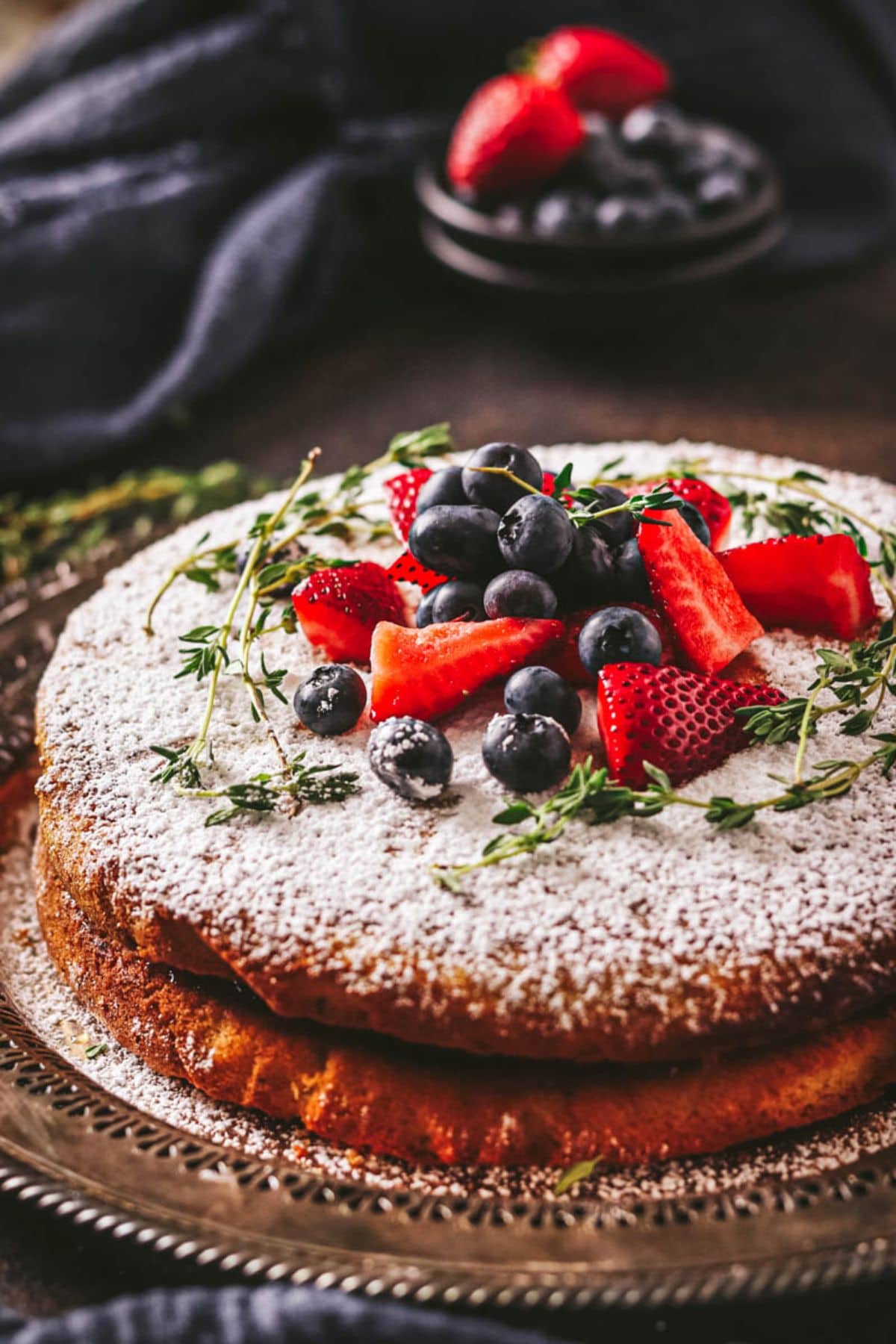 A round gluten free lemon cake dusted with powdered sugar, topped with fresh strawberries, blueberries, and green herbs, sits on an ornate silver tray. A small plate of fruit is in the blurred background.