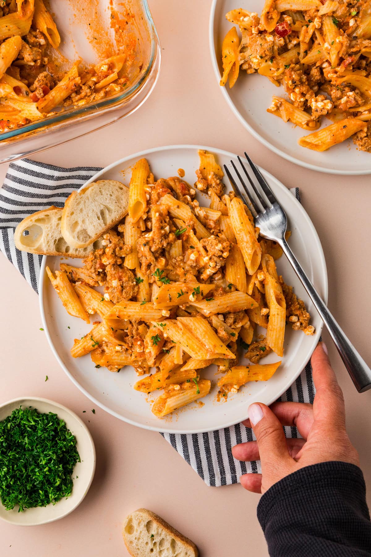 A hand reaches for a plate of 5 ingredient baked ziti with tomato sauce and cheese, garnished with herbs, beside a fork, bread slices, and a small bowl of chopped parsley on a striped napkin.