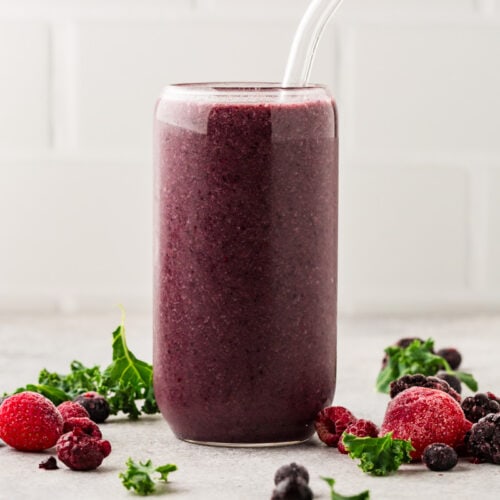 berry kale smoothie in a tall glass with frozen berries around it on a table.