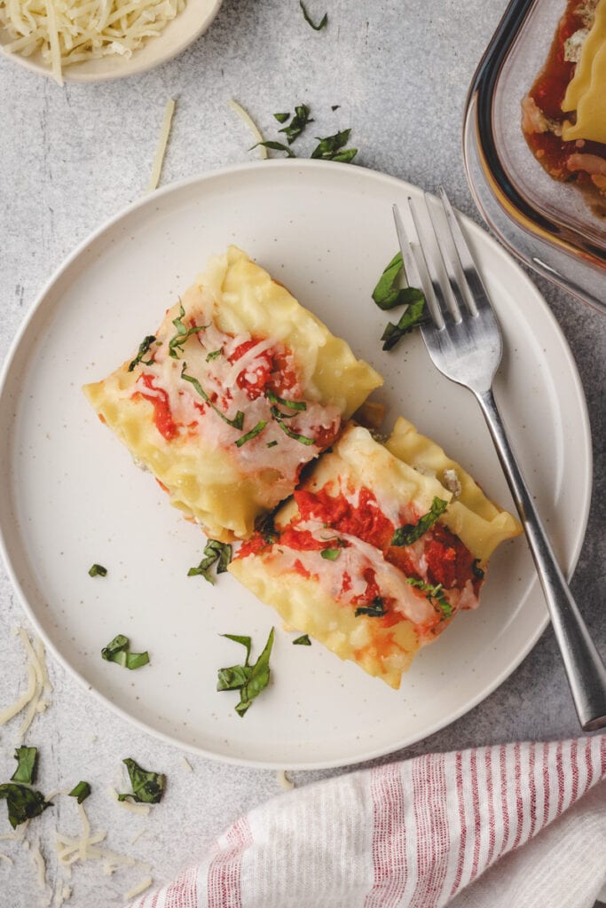 A plate with two turkey lasagna roll ups topped with tomato sauce, melted cheese, and chopped basil sits next to a fork. Shredded cheese and fresh basil are scattered nearby.