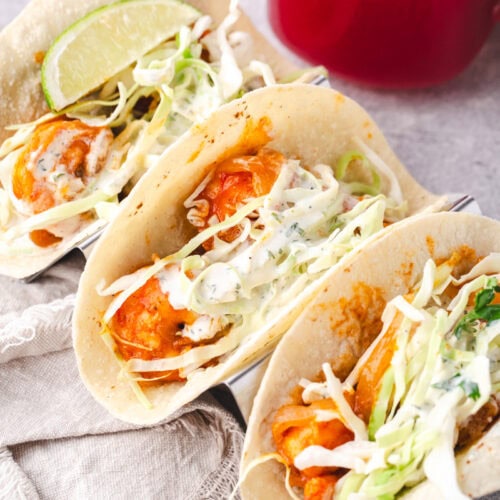 3 shrimp tacos in a taco holder with a strawberry drink in the background and a napkin.