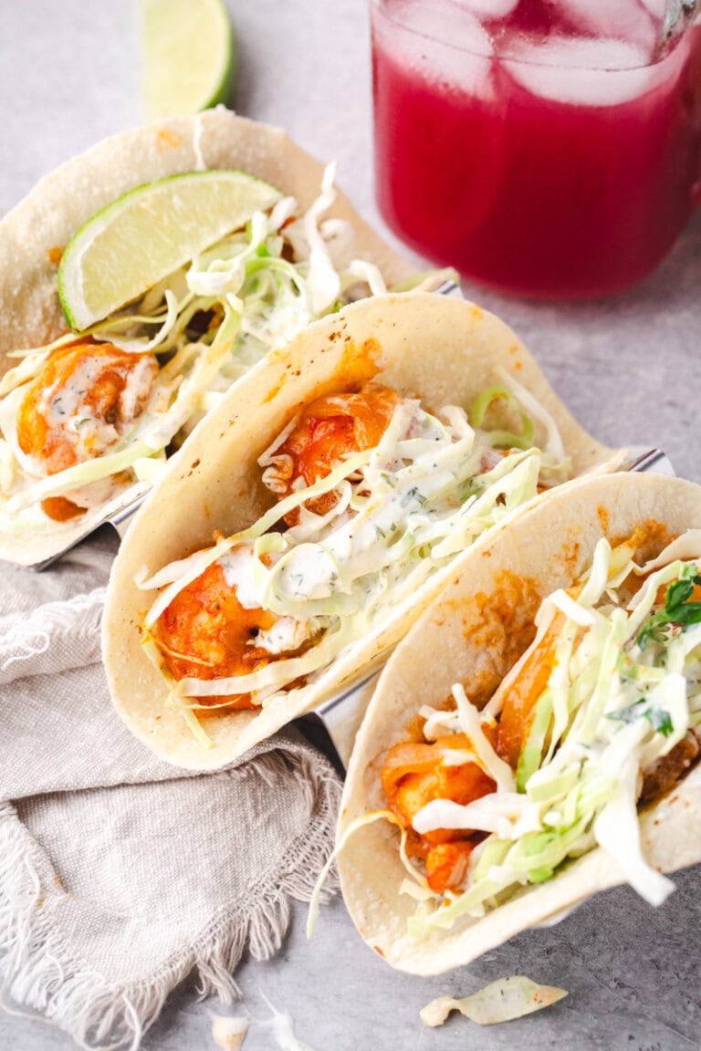 3 shrimp tacos in a taco holder with a strawberry drink in the background and a napkin.
