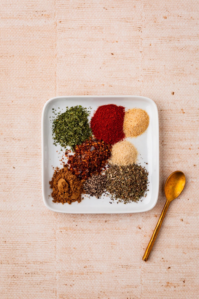 A white square plate displays an assortment of six colorful ground spices arranged in a circle to make all purpose seasoning. A small gold spoon rests beside the plate on a textured beige surface.