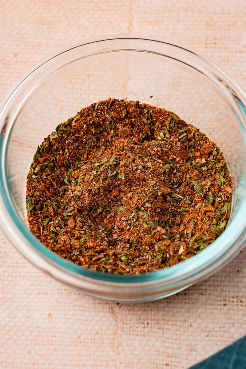 A clear glass bowl filled with all purpose seasoning , ncluding green and brown hues, sits on a textured beige surface. A portion of a dark blue cloth is visible at the bottom right corner.