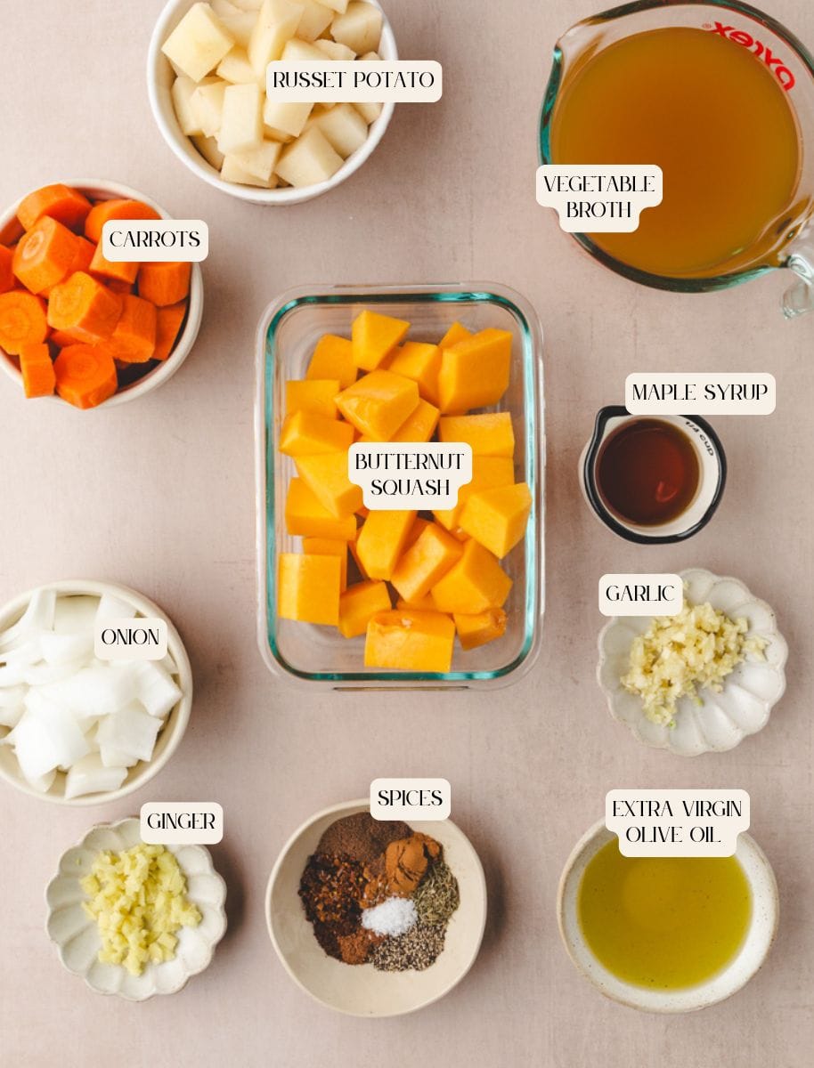 Ingredients for a hearty butternut squash and carrot soup are arranged on a beige surface: cubed russet potatoes, sliced carrots, cubed butternut squash, vegetable broth in a measuring cup, maple syrup, minced garlic, diced onion, ginger, spices, and olive oil.