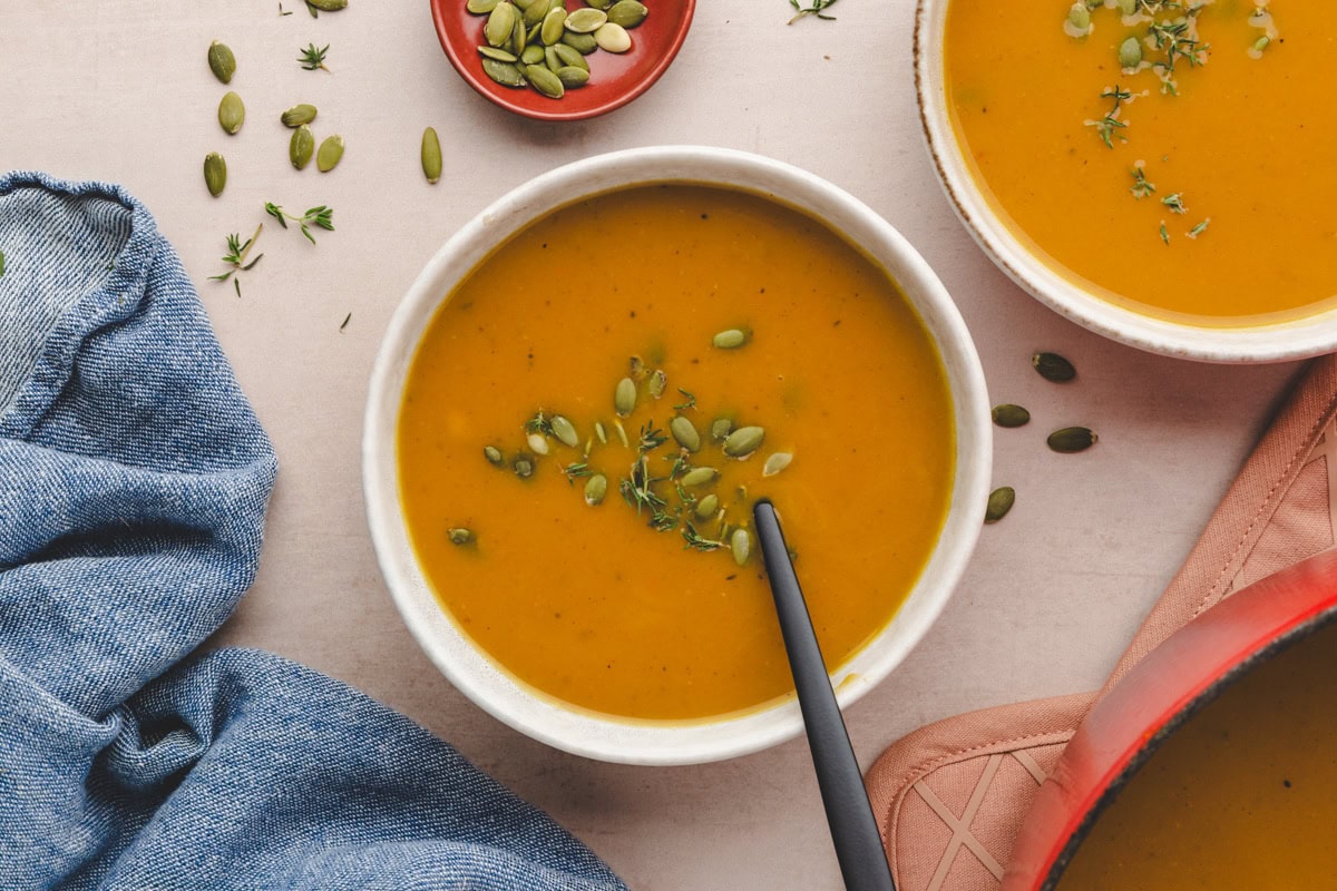 A bowl of pumpkin soup, with a hint of butternut squash, garnished with pumpkin seeds and thyme. A spoon rests inside the bowl, placed on a light-colored surface. Nearby, a denim cloth and a small plate of pumpkin seeds complement another soup bowl.